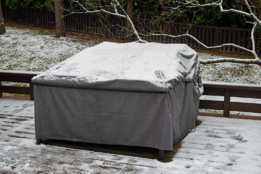 Outdoor patio furniture covered with a protective cover during winter snowfall on a deck.